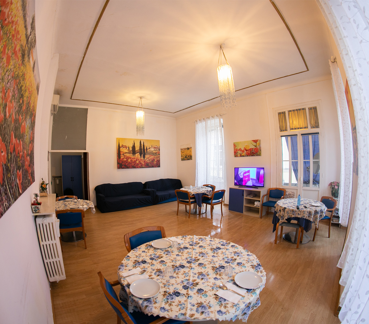 Sala comune con tavoli da pranzo, divano blu e grandi finestre – Residenza Anni Sereni